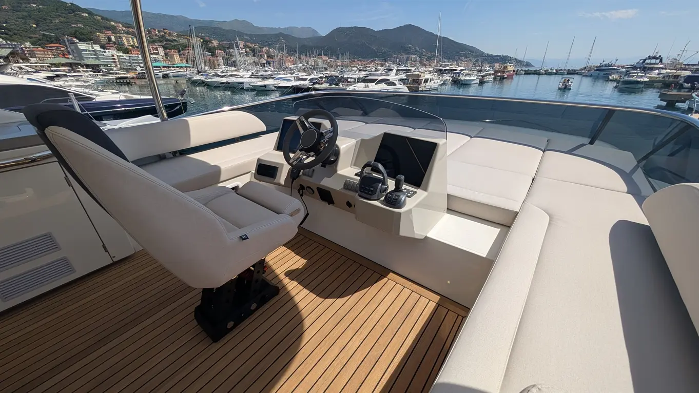 Luxury yacht cockpit with a steering wheel, two displays, and plush seating, overlooking a marina filled with boats.